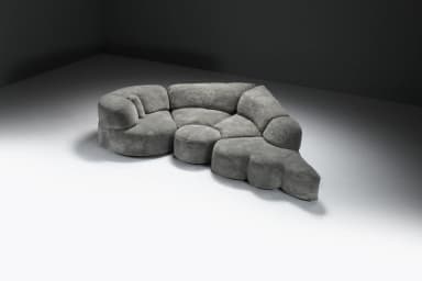 A large, multi-part sectional sofa featuring soft, organic, rounded forms, upholstered in a textured grey fabric. The sofa comprises several modules including corner pieces, armless central sections, and an end module that functions as an extended footrest or chaise. The overall design emphasizes comfort and a flowing, contemporary aesthetic. — 3D model