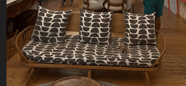 A mid-century modern style settee featuring a solid wood frame with a distinctive spindle back and integrated armrests. It is equipped with two seat cushions and three throw pillows, all upholstered in a geometric grey and white patterned fabric. The frame exhibits a natural wood finish, and the legs are tapered and splayed for stability and aesthetic appeal. The overall design suggests a blend of Scandinavian and mid-century modern influences. — 3D model