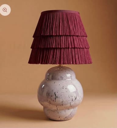 A table lamp featuring a rounded, bulbous ceramic base with a speckled finish and a tiered, pleated fabric shade. The base is a light, glossy hue with dark speckles, while the shade is a deep maroon or burgundy color, composed of several pleated fabric tiers that create a ruffled effect. — 3D model