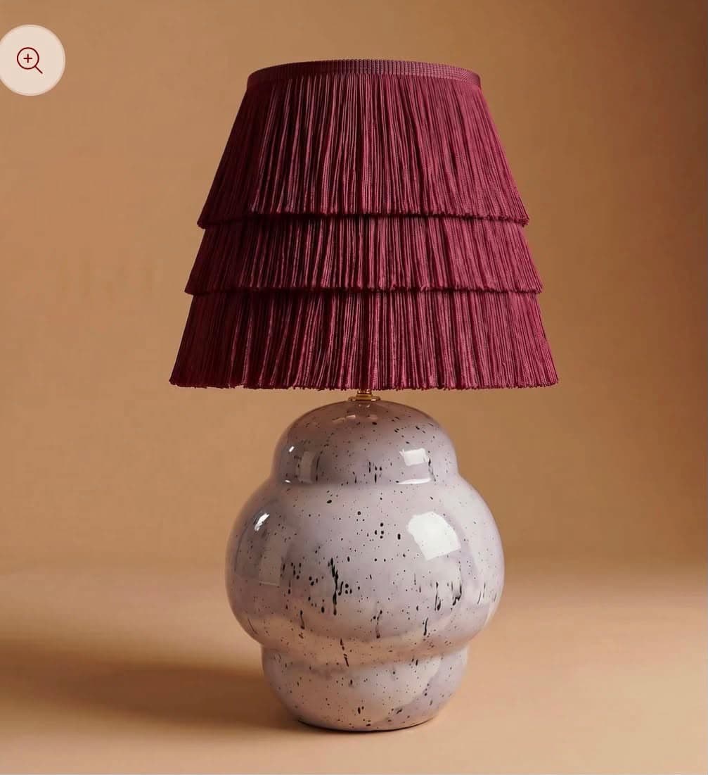 A table lamp featuring a rounded, bulbous ceramic base with a speckled finish and a tiered, pleated fabric shade. The base is a light, glossy hue with dark speckles, while the shade is a deep maroon or burgundy color, composed of several pleated fabric tiers that create a ruffled effect. — 3D model