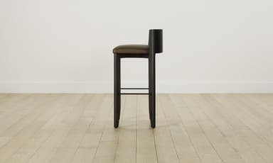A modern bar stool featuring a dark stained wooden frame and a padded, upholstered seat with a low backrest. The stool has four sturdy, slightly tapered legs and an integrated metal footrest. — 3D model