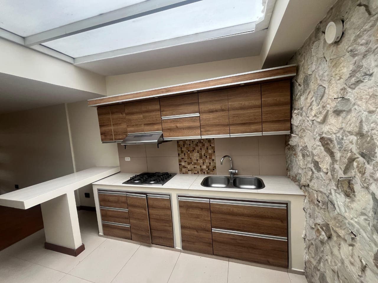 A compact, modular kitchen unit featuring light brown wood-grain laminate cabinets, white laminate countertops, a double-basin stainless steel sink, a four-burner gas stovetop, and a cantilevered white dining counter extension. An integrated exhaust hood and a mosaic-tiled backsplash are also visible. — 3D model