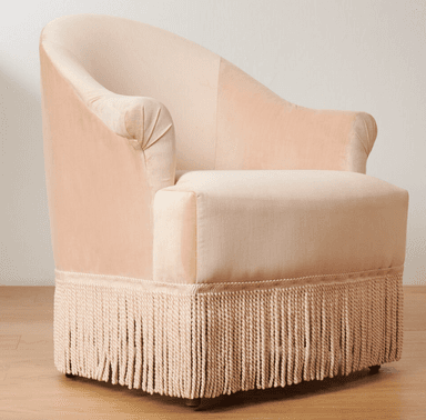 A light pink velvet armchair with a rounded back and arms. It features decorative pleating on the armrests and a prominent fringe along the bottom base. The seat cushion is a lighter, textured fabric. The chair has short, dark wooden legs visible at the very bottom. — 3D model