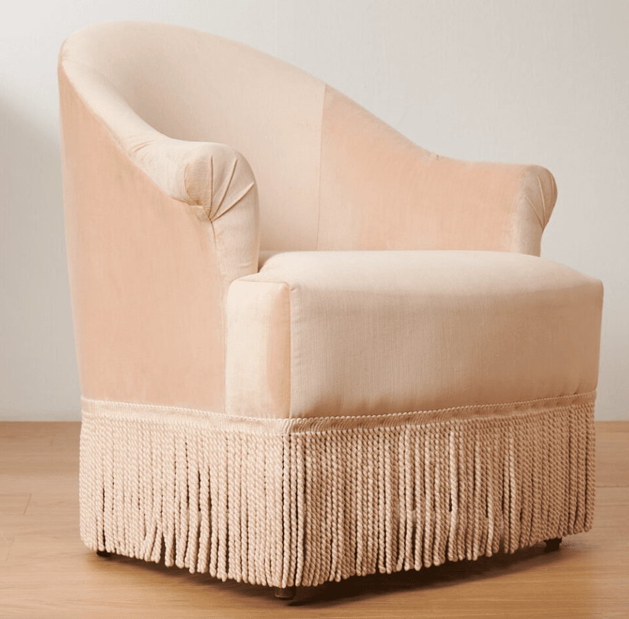 A light pink velvet armchair with a rounded back and arms. It features decorative pleating on the armrests and a prominent fringe along the bottom base. The seat cushion is a lighter, textured fabric. The chair has short, dark wooden legs visible at the very bottom. — 3D model