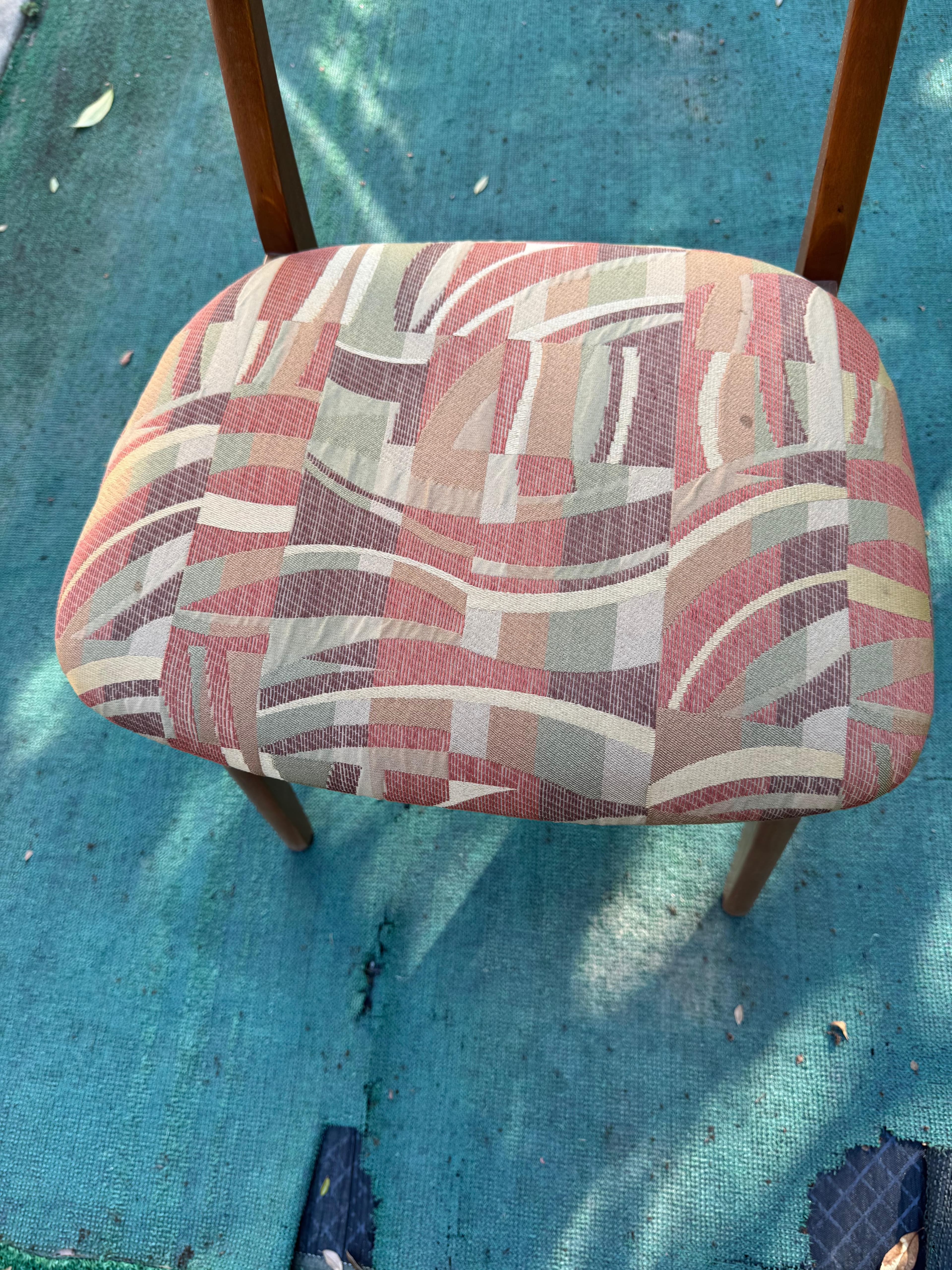 A close-up, overhead view of the padded seat and upper back support of an upholstered chair. The seat is covered in a multi-colored woven fabric featuring an abstract geometric pattern of wavy lines and rectangular blocks in shades of red, pink, beige, light green, and brown. Portions of the chair's wooden frame, including parts of the legs and back support, are visible, suggesting a medium-toned wood construction. The chair is positioned on a worn, teal-colored outdoor carpet or mat. — 3D model