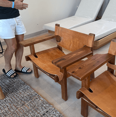 A craftsman-style lounge chair featuring a sturdy, light-toned wooden frame with a slung natural leather seat and backrest. The backrest includes distinctive circular cutouts, and wide wooden armrests are integrated into the robust frame. — 3D model