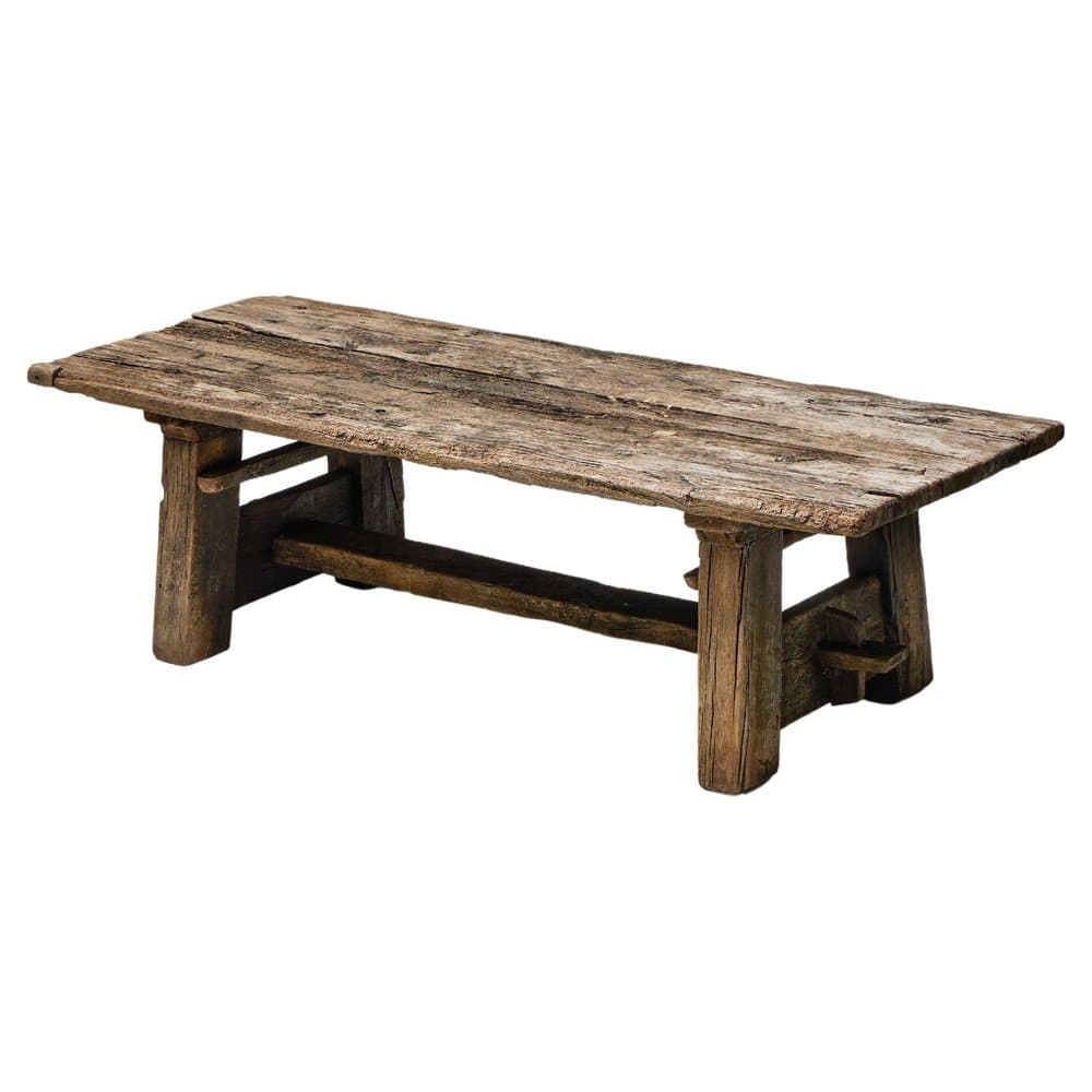 A rustic, distressed wooden bench featuring a thick, rectangular slab top and a robust trestle-style base. The wood exhibits significant weathering, including cracks, discoloration, and an uneven texture, suggesting age or intentional distressing. The construction appears simple and sturdy, with thick leg posts and a substantial crossbar providing support. — 3D model
