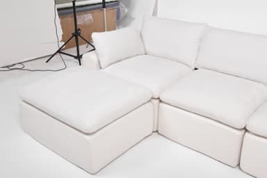 A modular sectional sofa with an attached ottoman. The sofa features deep, plush cushions and a low profile. It is upholstered in a textured white fabric. The configuration shown includes a corner section, two seat cushions, and an ottoman. The design is contemporary and emphasizes comfort. — 3D model