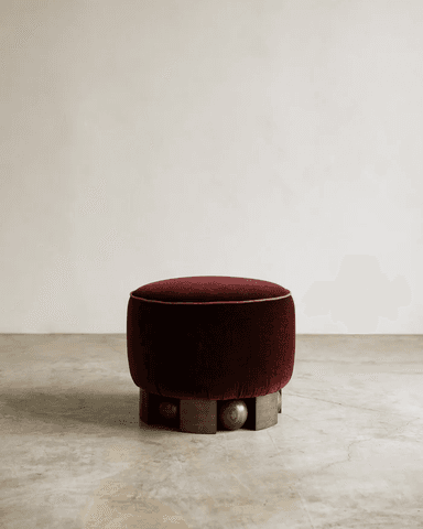 A round, upholstered ottoman with a deep red velvet fabric. The ottoman is characterized by a decorative piping along its top edge and a unique base constructed from dark wood elements, including carved spherical shapes and angular blocks arranged in a tiered pattern. The base provides a contrast to the soft upholstery of the seat. — 3D model