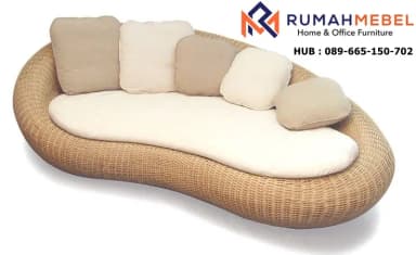 A contemporary outdoor daybed featuring an organic, kidney-bean shape. The base is constructed from woven synthetic rattan or natural wicker in a light brown hue. It is topped with a custom-fitted, off-white or cream-colored mattress, providing a comfortable lounging surface. Several loose throw pillows in complementary beige and cream tones are arranged along one curved edge, serving as a flexible backrest. The design suggests a relaxed, resort-style aesthetic, suitable for outdoor living spaces. — 3D model