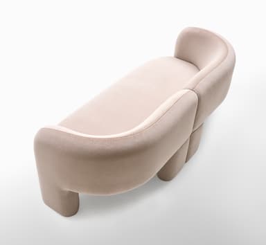 A contemporary loveseat upholstered in a light, neutral fabric, featuring a distinctive curved backrest and integrated rounded armrests. The piece has a monolithic appearance with no visible legs, suggesting a continuous, low-profile base. The design emphasizes soft lines and a plush, inviting form, suitable for modern interior settings. The fabric appears to have a subtle texture. — 3D model