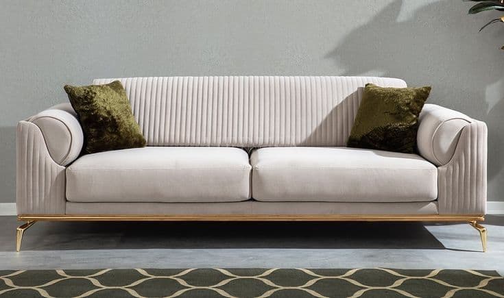 A modern three-seater sofa featuring plush light-colored upholstery, likely cream or off-white, with distinctive channel tufting on the armrests and backrest. The sofa is supported by a sleek, polished brass-finished metal frame that forms the base and extends into four tapered legs. Two olive green throw pillows are included. — 3D model