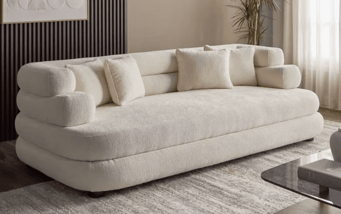 A modern sofa upholstered in a light cream bouclé fabric, featuring a distinctive low-profile design with multiple stacked, rounded sections forming the backrest and arms. The sofa includes several matching bouclé throw pillows and rests on discreet, dark cylindrical feet. — 3D model