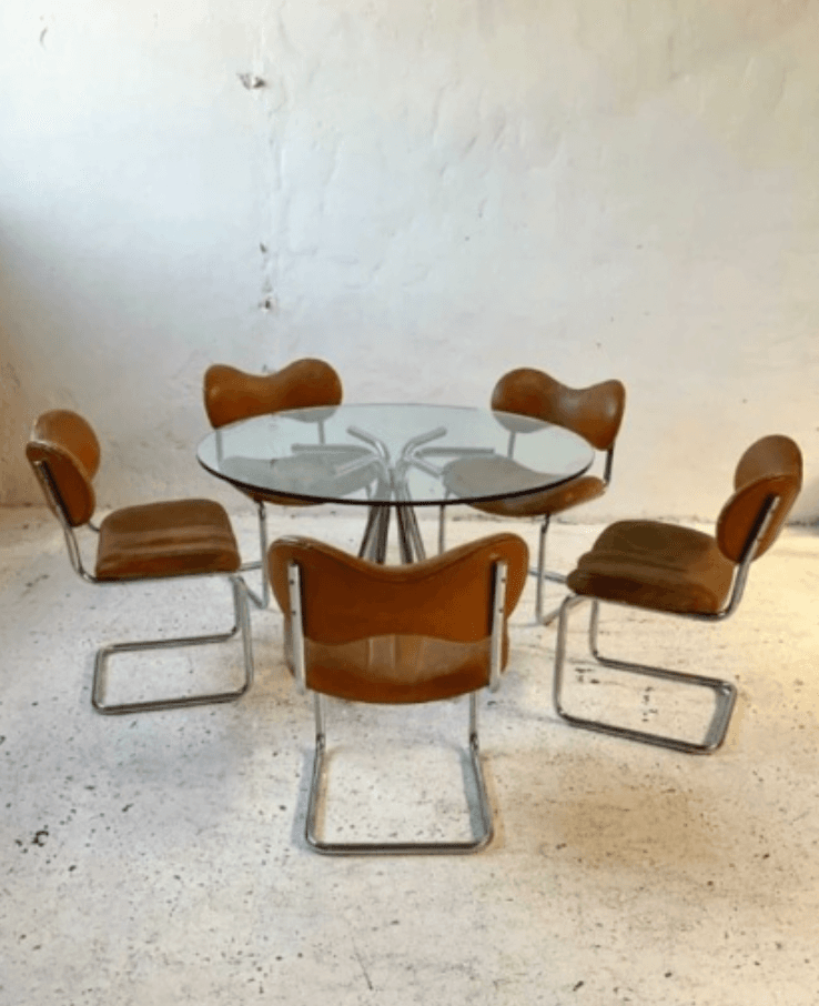 A round glass-top dining table with a chrome base, accompanied by five matching cantilever chairs. The chairs have chrome frames and brown leather upholstery on the seats and backs. The table top appears to be circular with a clear glass surface supported by a central pedestal base with radiating arms. — 3D model