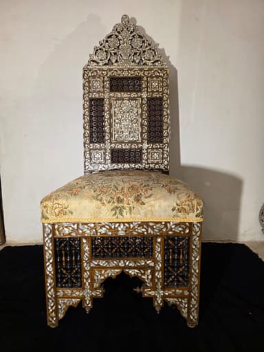 A highly ornate accent chair featuring intricate marquetry and inlay work. The chair has a tall, elaborately carved back with floral motifs and geometric patterns. The seat is upholstered in a light-colored fabric with a floral pattern. The base of the chair, including the legs and the area beneath the seat, is constructed from dark wood and heavily decorated with mother-of-pearl inlay, creating floral and geometric designs. The legs have an arched structure. The overall construction suggests a piece of antique or artisan craftsmanship. — 3D model