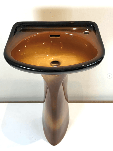 A contemporary pedestal sink featuring a striking ombré finish that transitions from dark black at the rim to a warm, glossy brown in the basin and down the pedestal. The basin has an elliptical shape with a single faucet hole and an integrated overflow slot. The pedestal base tapers elegantly from the basin to the floor, contributing to a sleek and modern aesthetic. — 3D model