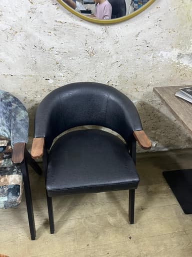 A modern dining chair featuring a barrel-shaped backrest and seat upholstered in black faux leather, complemented by dark wooden armrest caps and a matte black metal frame. — 3D model