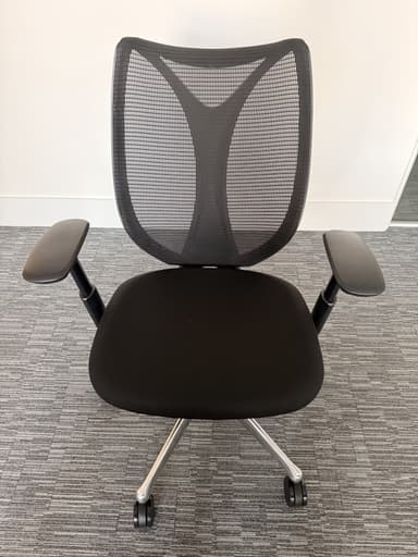 A modern ergonomic office chair is depicted from a frontal, slightly elevated perspective, set against a white wall and light grey carpeted floor. The chair features a black mesh backrest with a pronounced lumbar support curve, a black fabric-upholstered seat, and adjustable black plastic armrests. It is supported by a polished metallic five-star swivel base equipped with five black caster wheels. — 3D model