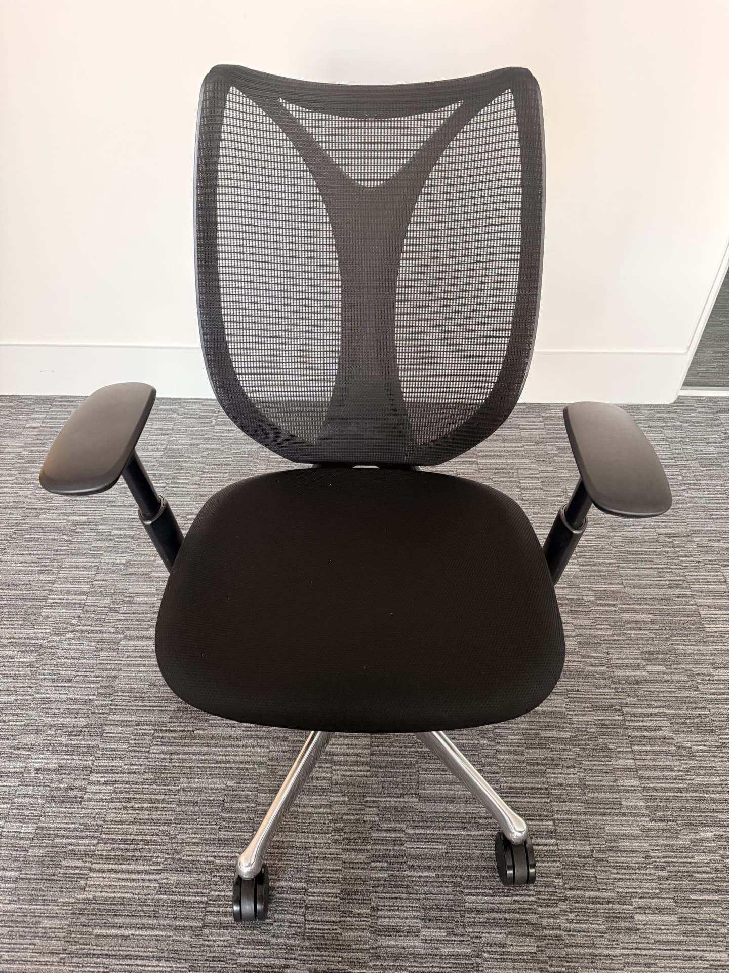 A modern ergonomic office chair is depicted from a frontal, slightly elevated perspective, set against a white wall and light grey carpeted floor. The chair features a black mesh backrest with a pronounced lumbar support curve, a black fabric-upholstered seat, and adjustable black plastic armrests. It is supported by a polished metallic five-star swivel base equipped with five black caster wheels. — 3D model