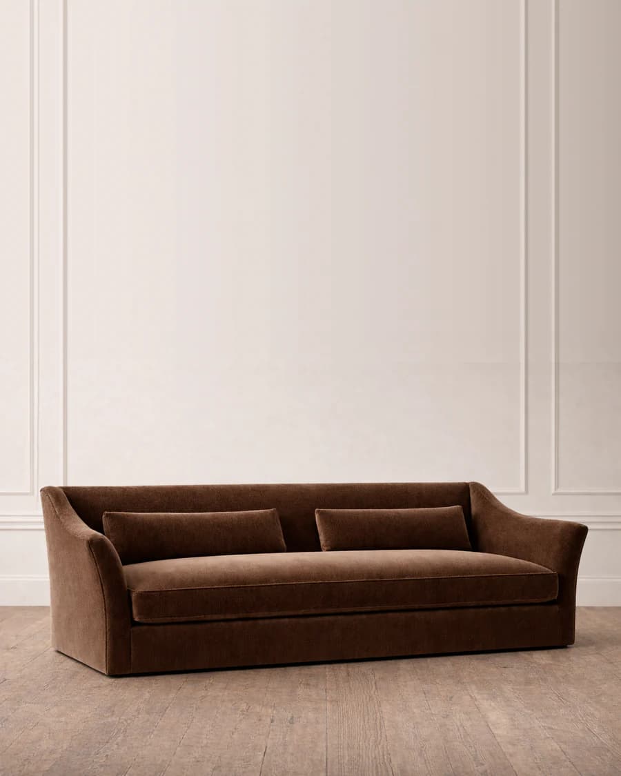 A plush, upholstered sofa with a warm brown velvet fabric. It features a clean, tailored design with a low profile and gently sloping arms that curve upwards. The sofa has a single seat cushion and a back cushion that appears to be integrated or very closely fitted. Two rectangular throw pillows in the same fabric are placed against the back. The base of the sofa is a solid, squared-off structure. It rests on a wooden floor with visible planking. — 3D model