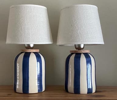 A table lamp featuring a cylindrical ceramic base with a bold blue and white vertical stripe pattern, topped with a tapered drum fabric lampshade in a neutral beige or cream color. The lamp has a clean, rendered appearance. — 3D model