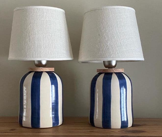 A table lamp featuring a cylindrical ceramic base with a bold blue and white vertical stripe pattern, topped with a tapered drum fabric lampshade in a neutral beige or cream color. The lamp has a clean, rendered appearance. — 3D model