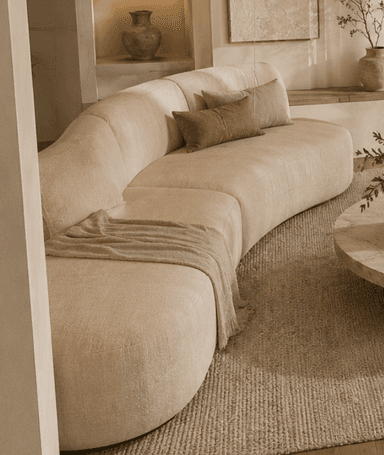 A large, modern, curved sectional sofa upholstered in a light beige, textured fabric. It features a continuous, gently curved backrest and armrest on one end, and an open end on the other, suggesting a modular design. The base is concealed, giving it a low-profile appearance. Several matching and contrasting throw pillows are placed on the seats, along with a light brown throw blanket. — 3D model