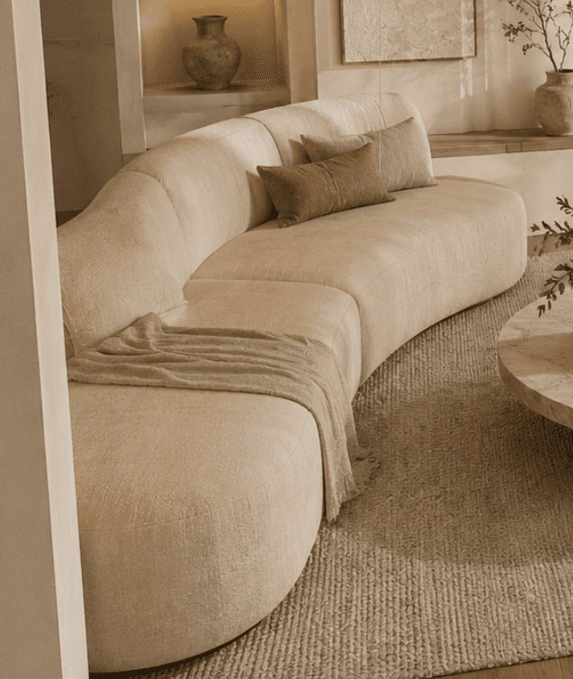 A large, modern, curved sectional sofa upholstered in a light beige, textured fabric. It features a continuous, gently curved backrest and armrest on one end, and an open end on the other, suggesting a modular design. The base is concealed, giving it a low-profile appearance. Several matching and contrasting throw pillows are placed on the seats, along with a light brown throw blanket. — 3D model