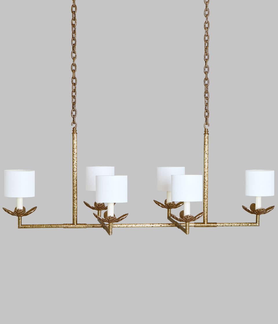 A linear chandelier suspended by two chains. The fixture has a horizontal bar with six lights, each featuring a white cylindrical lampshade supported by a decorative metal arm with leaf-like accents. The metal frame has a distressed gold finish. — 3D model