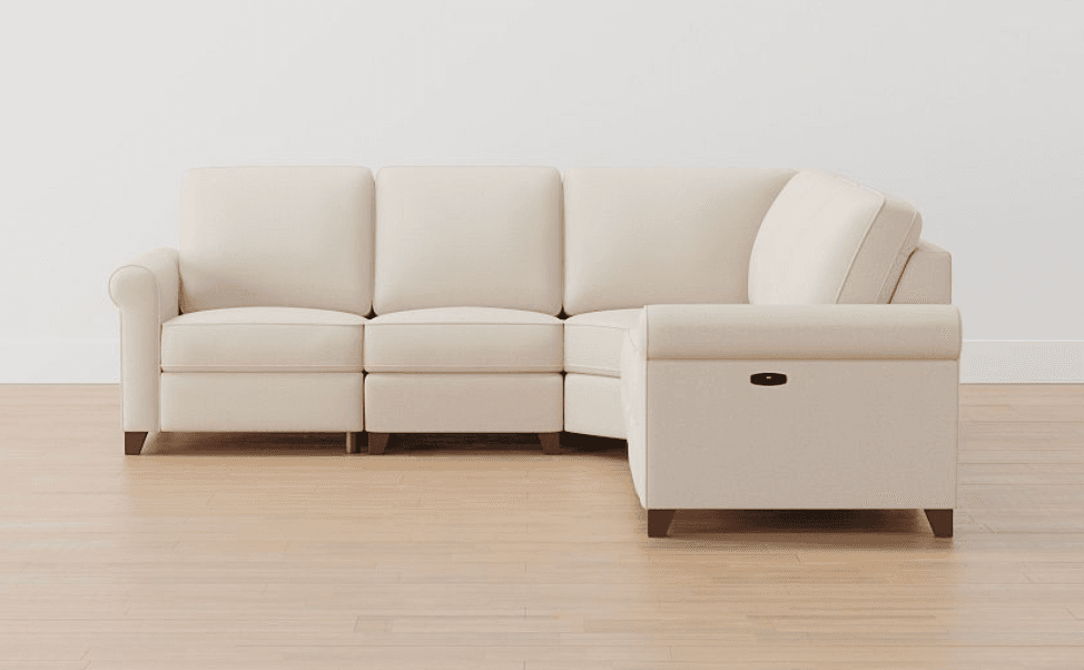A contemporary L-shaped sectional sofa upholstered in light cream fabric, featuring rolled armrests and multiple seat and back cushions. It is composed of several modules, including an L-corner piece and end sections that have integrated reclining functionality, indicated by a black lever on the armrest. The sofa is supported by short, tapered dark wood legs. — 3D model