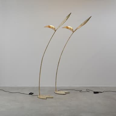 A tall, slender floor lamp with an organic, botanical design, featuring a distressed gold or bronze finish. The lamp has a round, flat base from which a curved stem extends upwards, culminating in two leaf-shaped elements that house the light sources. A black power cord with an inline foot switch extends from the base. — 3D model