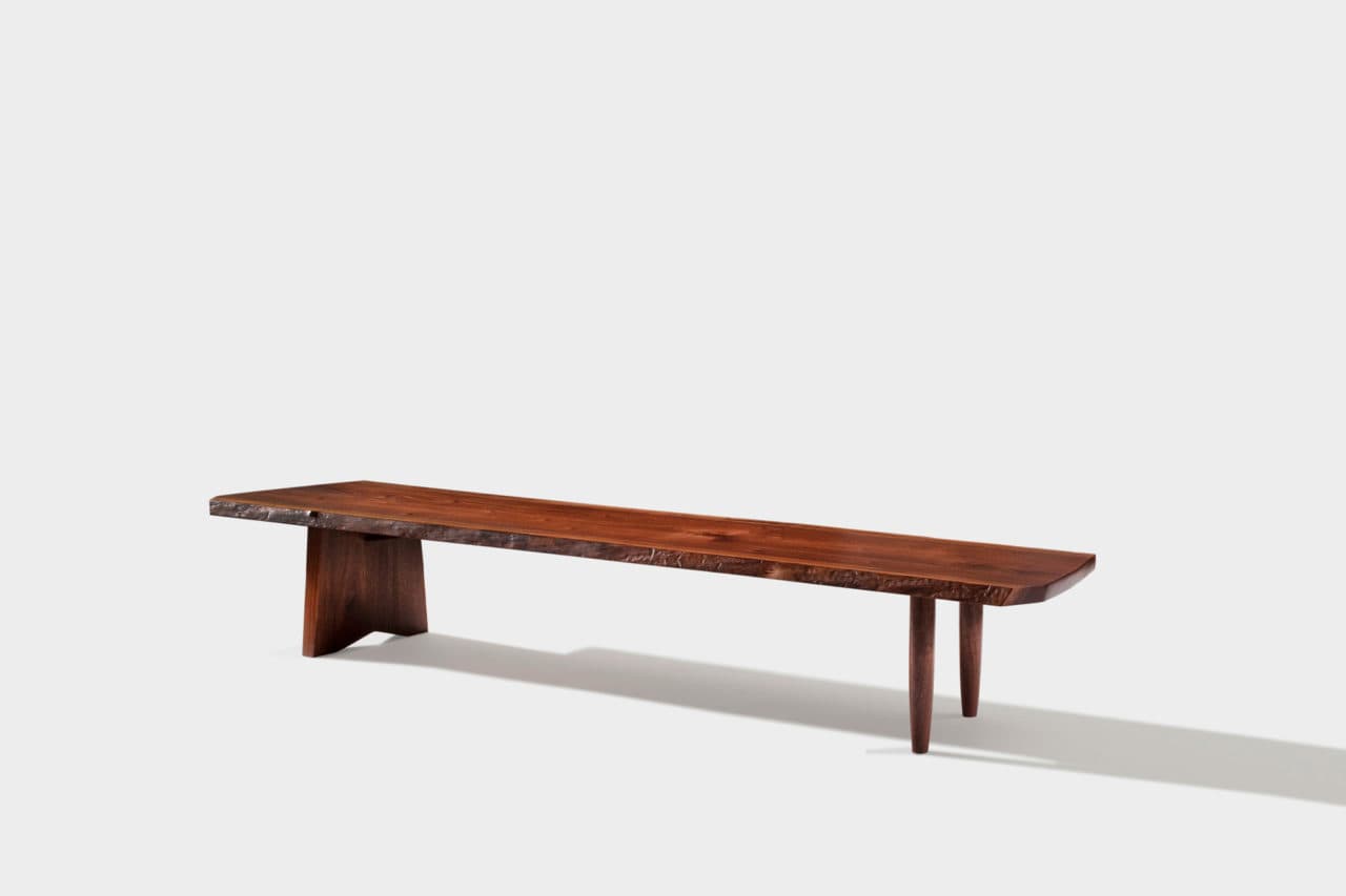 A long, narrow wooden bench with a live edge on one side and a finished surface. It is supported by two distinct leg structures: one is a solid, angled piece on the left, and the other consists of three tapered legs on the right. The wood exhibits a rich, dark brown color with visible grain patterns. — 3D model