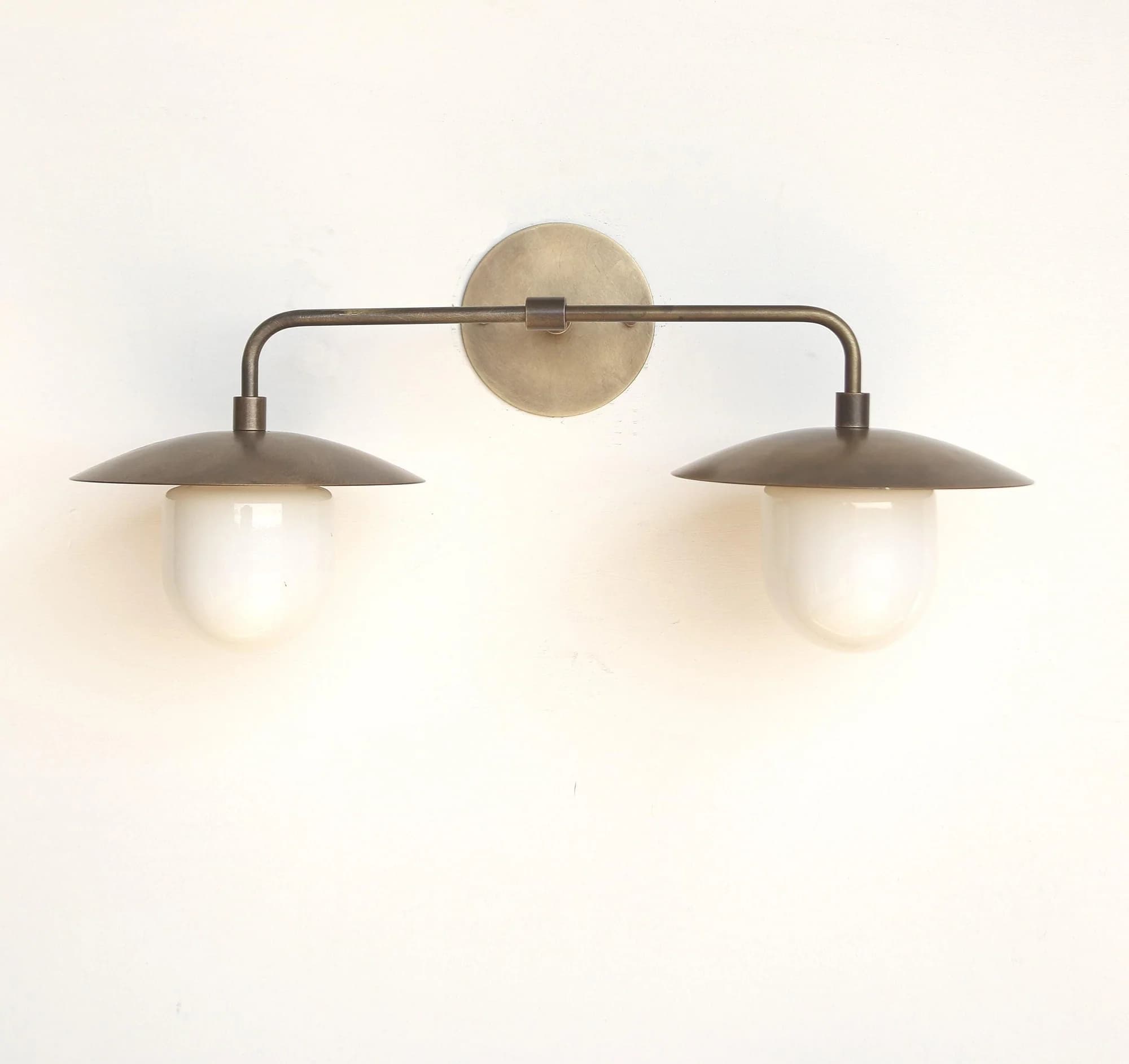 A double arm wall sconce featuring an aged brass or bronze finish. It consists of a circular backplate from which a horizontal arm extends. This arm then branches into two downward-curving arms, each holding a light source. Each light source is composed of a disc-shaped metal shade resting above a frosted white glass globe diffuser. — 3D model