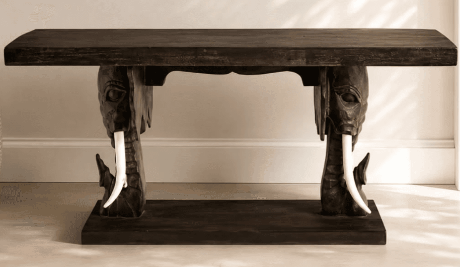 A dark wooden console table featuring intricately carved elephant heads as supports, complete with white tusks, connecting the tabletop to a lower base. — 3D model