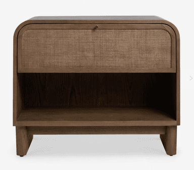 A contemporary nightstand featuring a dark-stained wooden frame with gently rounded top edges. It includes a single drawer with a woven cane front and a small brass knob, positioned above an open lower storage shelf. The base is a solid, plinth-like structure with subtle, short legs. — 3D model