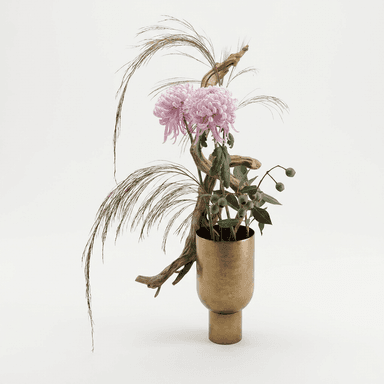 A sophisticated ikebana-inspired floral arrangement presented in a modern brass vase. The arrangement features large, pale pink chrysanthemums, delicate feathery dried grasses, and sprigs of eucalyptus with seed pods, all artfully interwoven with a natural driftwood branch. The vase itself is a cylindrical brass vessel with a tapering neck and a stout circular base, exhibiting a brushed or matte finish. — 3D model