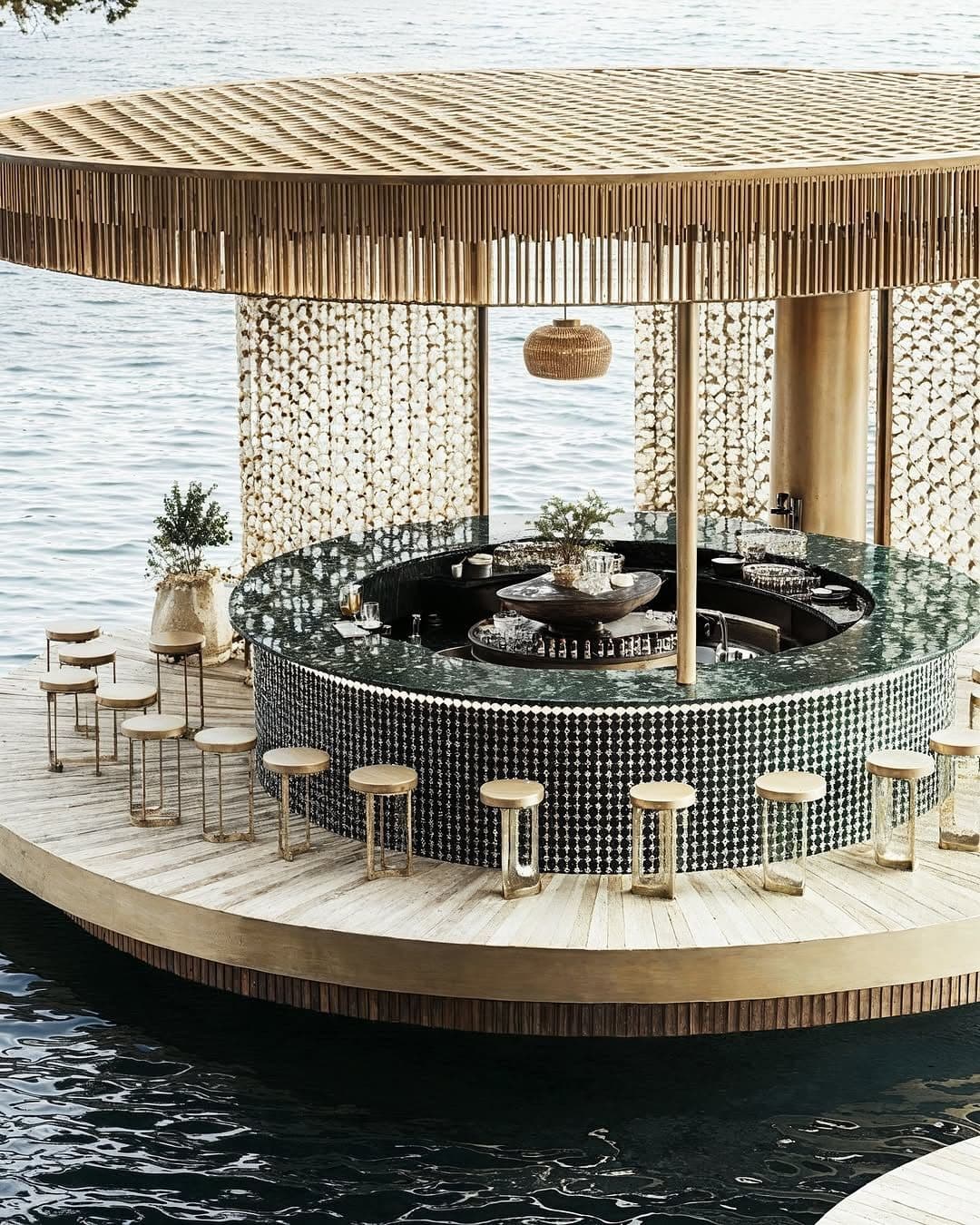A circular bar with a green speckled countertop and a patterned facade. It features a central raised display area with decorative items and glassware. Several bar stools with light wood seats and metal frames are positioned around the perimeter. The background includes decorative wall panels and a hanging light fixture. — 3D model