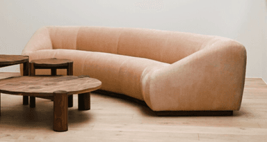 A contemporary, generously curved upholstered sofa in a light, neutral beige tone. The sofa features a continuous, rounded form with no visible legs, giving it a low-profile and substantial presence. The backrest and seat are seamlessly integrated, suggesting a cohesive, single-piece construction. The upholstery appears to be a textured fabric, possibly boucle or a fine-grained velvet, with subtle vertical seams segmenting the back and seat portions. Its design is minimalist, focusing on organic curves and comfort. — 3D model