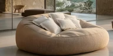 A large, round, bean bag-style seating unit upholstered in a textured, light beige fabric. It features a soft, unstructured form and is accompanied by several small, off-white throw pillows, some with fringed edges, arranged on its surface. The overall appearance suggests a comfortable and casual seating option. — 3D model