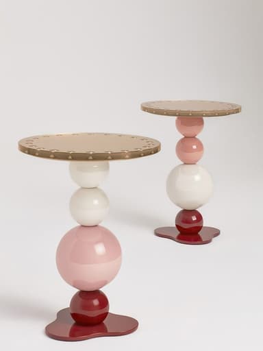 A postmodern accent table featuring a distinctive stacked spherical base in various colors, topped with a circular metallic surface. The base consists of a dark red organic-shaped foot, supporting a smaller dark red sphere, a larger light pink sphere, and a white sphere, arranged vertically. The top is a flat, round surface with a metallic gold or brass finish, detailed with a repeated pattern of small embossed or debossed elements around its edge. — 3D model