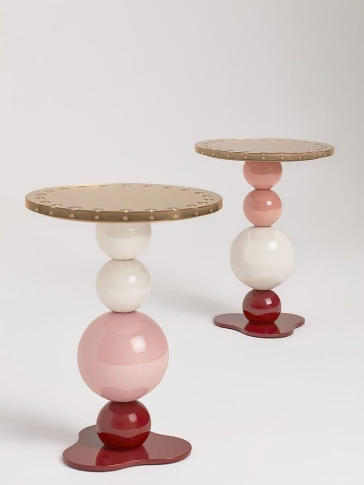 A postmodern accent table featuring a distinctive stacked spherical base in various colors, topped with a circular metallic surface. The base consists of a dark red organic-shaped foot, supporting a smaller dark red sphere, a larger light pink sphere, and a white sphere, arranged vertically. The top is a flat, round surface with a metallic gold or brass finish, detailed with a repeated pattern of small embossed or debossed elements around its edge. — 3D model