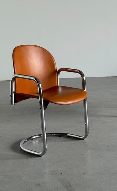 A cantilever armchair with a chrome-plated tubular steel frame and a brown leather seat and backrest. The frame has a continuous curve forming the legs, seat support, and armrests. The backrest is slightly curved and extends upwards, with a visible seam running along its edge. The seat is a flat plane that connects to the frame. The overall design suggests a mid-century modern or Bauhaus influence. — 3D model