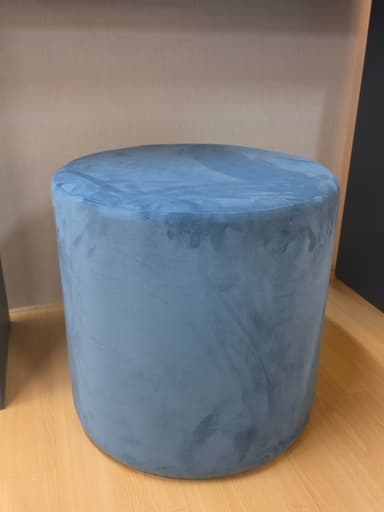 A cylindrical ottoman, fully upholstered in a plush, blue-gray velvet fabric. The ottoman presents a simple, unadorned form with no visible legs or distinct base, giving it a monolithic appearance. The velvet material shows varying tones due to light reflection on its pile, indicating its soft texture. — 3D model