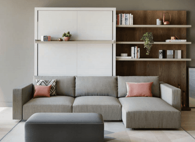 A sectional sofa with a chaise lounge extension and a matching ottoman. The sofa is upholstered in a light grey textured fabric and features clean lines and a low profile. It includes two decorative throw pillows: one solid pink and one with a geometric pattern in shades of grey and pink. The ottoman is rectangular and upholstered in a dark grey fabric with a subtle piped edge. — 3D model