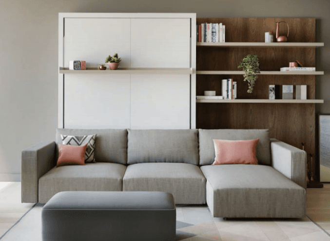 A sectional sofa with a chaise lounge extension and a matching ottoman. The sofa is upholstered in a light grey textured fabric and features clean lines and a low profile. It includes two decorative throw pillows: one solid pink and one with a geometric pattern in shades of grey and pink. The ottoman is rectangular and upholstered in a dark grey fabric with a subtle piped edge. — 3D model