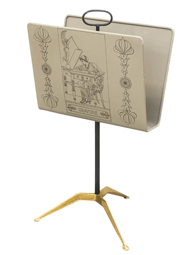 A freestanding magazine rack featuring a light-colored enameled metal trough adorned with black Art Deco-style line art illustrations. It is supported by a slender dark metal central pole and a distinctive three-legged brass base. A dark metal loop handle is integrated at the top for portability. — 3D model