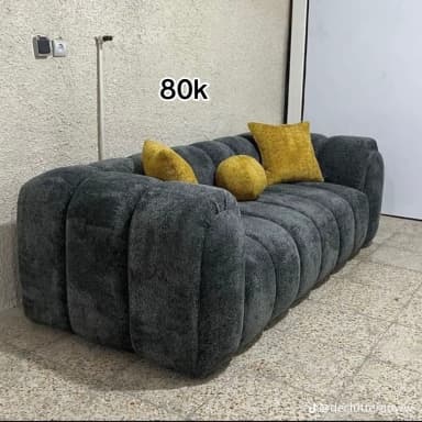 A modern sofa characterized by a distinctive ribbed or channel-tufted design in a dark grey, textured fabric. The sofa features a low-profile silhouette with wide, rounded armrests and backrest, creating a cohesive, sculptural form. It comes with three accent pillows in a mustard yellow hue: two square and one round, adding a pop of color to the monochromatic base. — 3D model