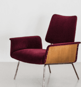 A modern armchair with a distinct mid-century aesthetic. It features a curved backrest and seat upholstered in a deep burgundy, possibly velvet or a similar textured fabric. The armrests are integrated into the main body of the chair. A prominent design element is the exposed wooden panel on the sides, appearing to be bent plywood, which contrasts with the upholstered sections. The chair is supported by slender, metallic legs that angle outwards. — 3D model
