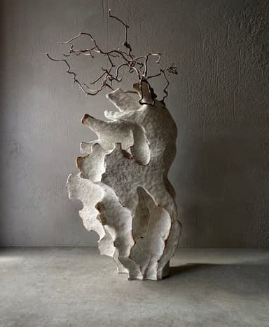 An abstract sculpture featuring a layered, biomorphic body with an organic, uneven surface texture, made from what appears to be unglazed ceramic or a similar composite material. From the top of the sculpture emerge several intricate, dried, and gnarled branch-like elements, creating a dynamic contrast between the solid base and the delicate, wiry extensions. The overall form suggests an organic growth or a natural geological formation. — 3D model