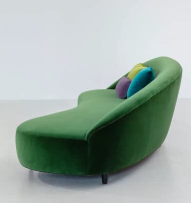 A modern chaise lounge upholstered in rich emerald green velvet, featuring a gracefully curved and ergonomic form. It has a low, integrated backrest that flows seamlessly into the seat and a footrest extension. The piece is supported by discreet, dark cylindrical legs, and three small, round accent pillows in lime green, purple, and teal are placed on the backrest. — 3D model